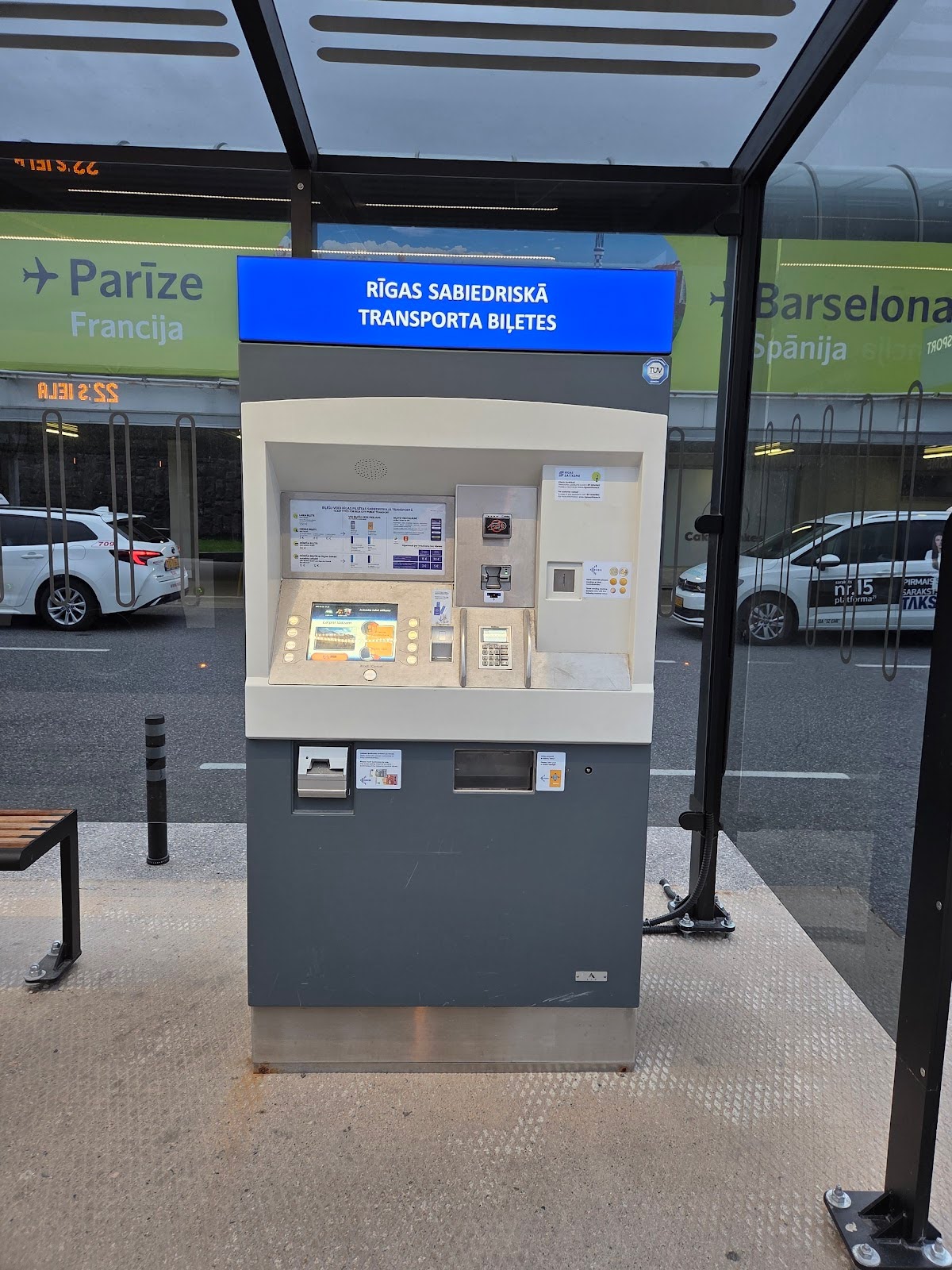 A public transport ticket machine in Riga, Latvia, at a covered outdoor stop with cars and city signs visible in the background.