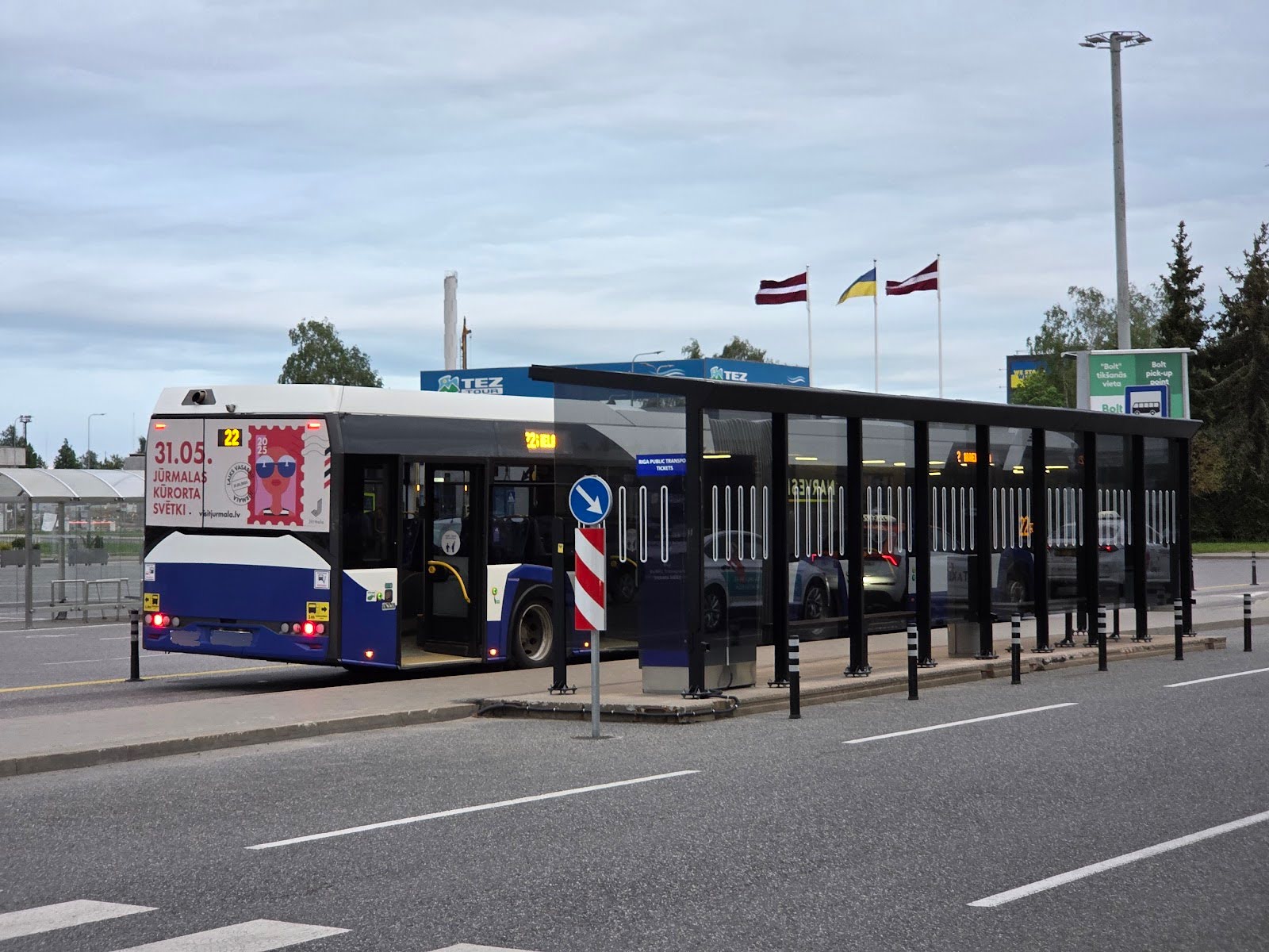 A city bus advertisement displays a colorful event poster at a modern bus stop, flanked by flags and nearby signage.
