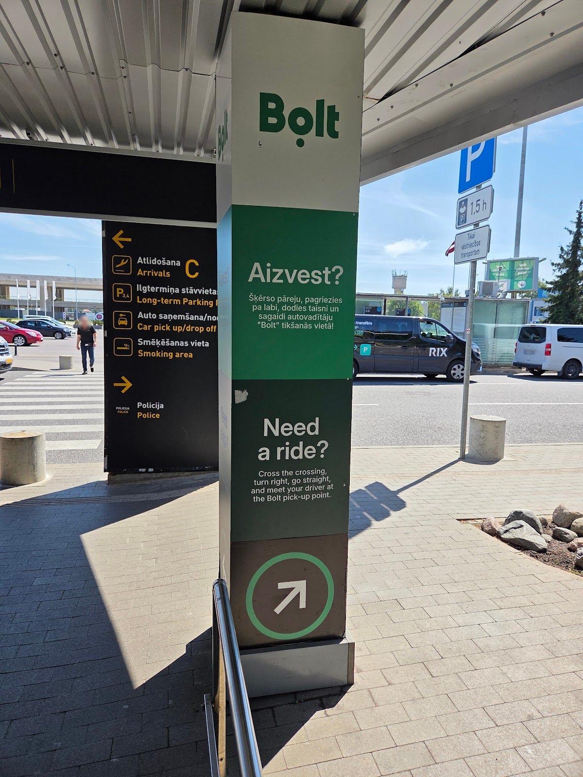 A green and white Bolt pillar sign at a curb shows directions for ride pick-up, with text in Latvian and English and an arrow pointing toward the Bolt pick-up site.
